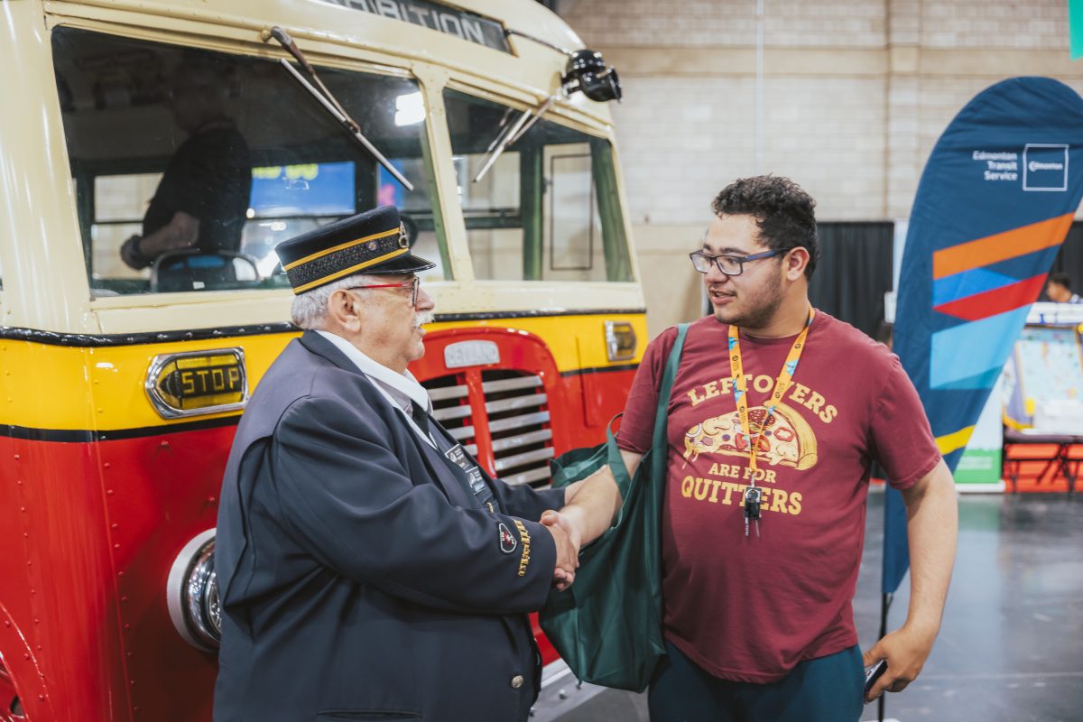 A man in an transit operator uniform smiles and shakes hands with a man in a tshirt in front of an exhibit with a vintage red, yellow and beige bus behind them.