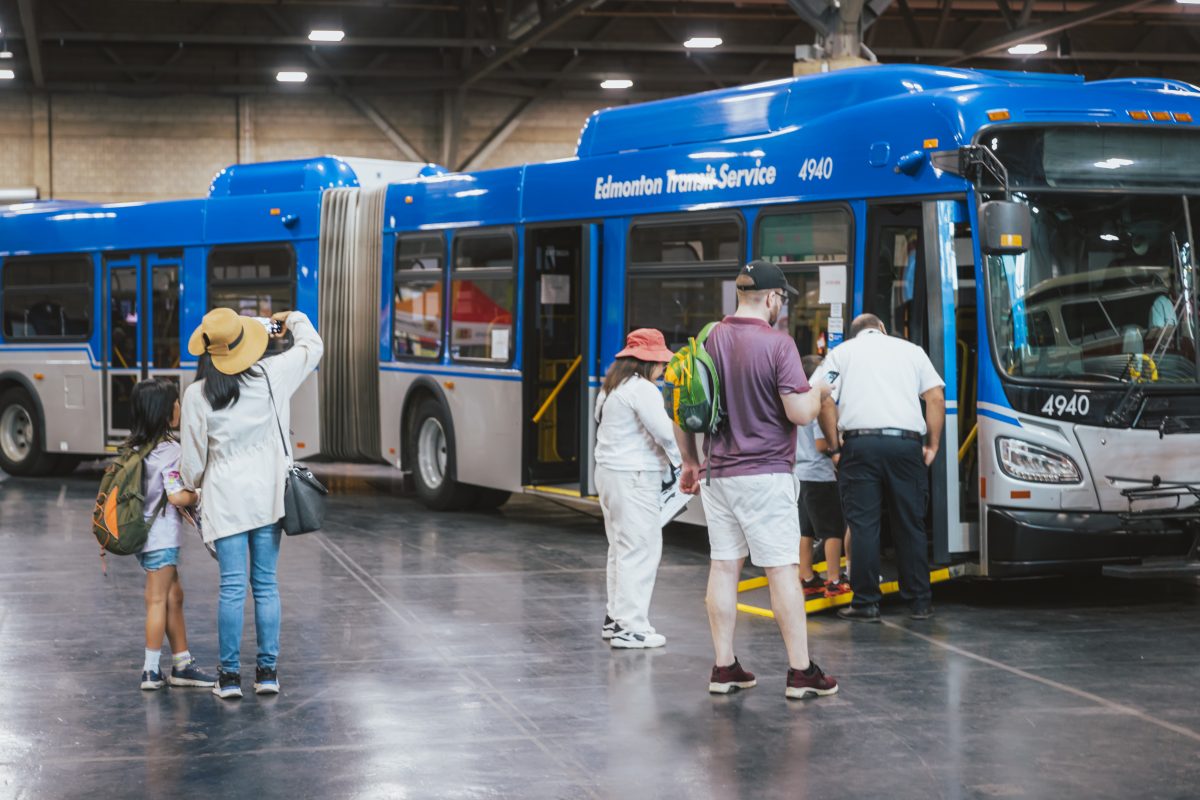 Multiple people stand in near a newer bluer and grey Edmonton Transit bus at an indoor exhibit.