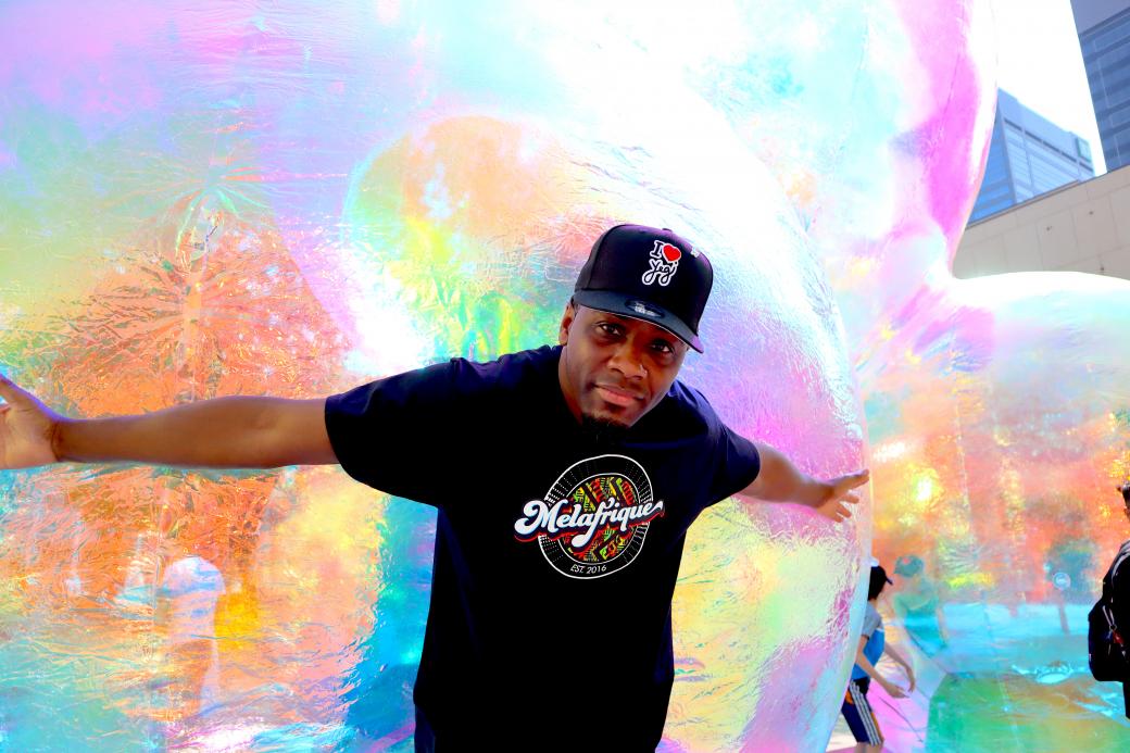 A black man facing the camera stands with his arms stretched around a giant artwork bubble. 