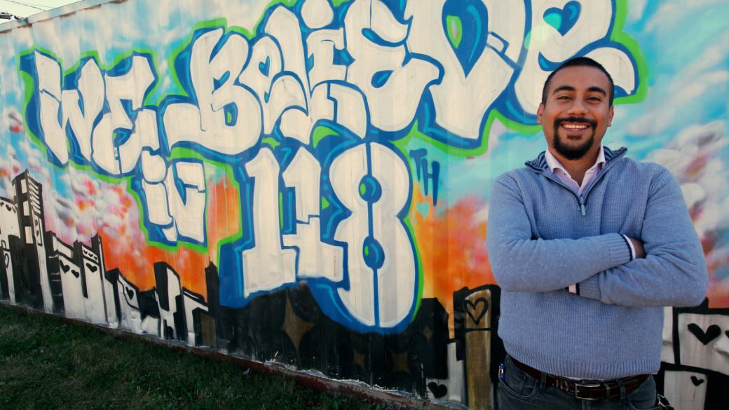 Man stands smiling with his arms crossed in front of a mural that says “We Believe in 118”.