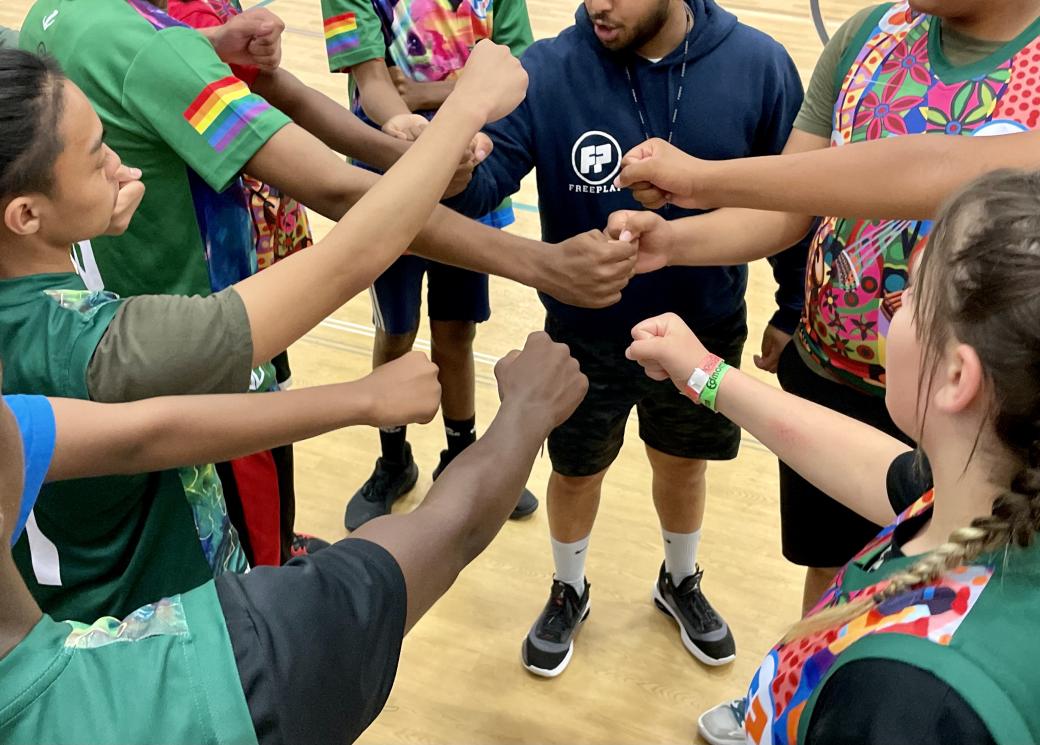 A group of basketball players and their coach extend their right arms as they huddle in a circle.