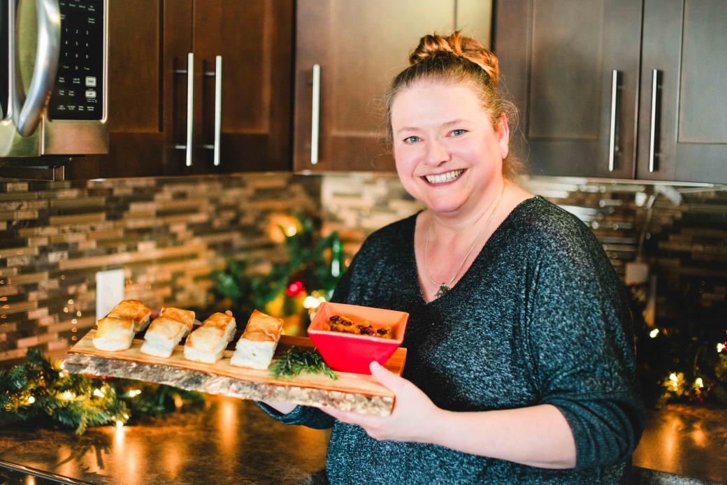  Kathy Neiman stands in her festive kitchen holding a platter of delicious tourtière puffs. A red dish of chutney complements the appetizers, and Christmas decorations twinkle in the background, creating a warm and inviting atmosphere.