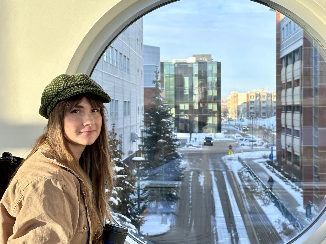 A woman with long hair and a green cap stands in front of a circular window, out of which four university buildings and a semi-snow covered road can be seen.