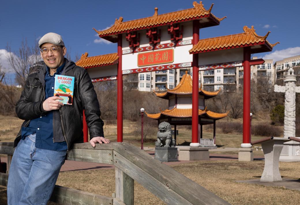 A man in a leather jacket, jeans and a cap holds up a book in front of a Chinese gate, lion statue and pagoda. 