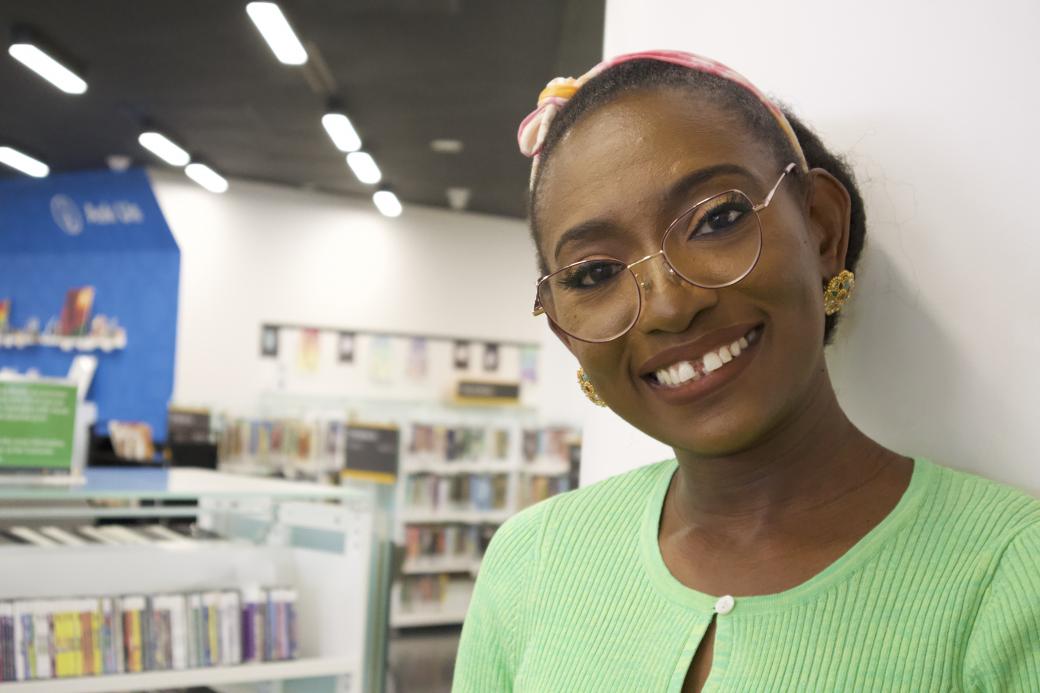 A Black woman with glasses smiles as she stands next to a pillar in a library with shelves of books in the background. 