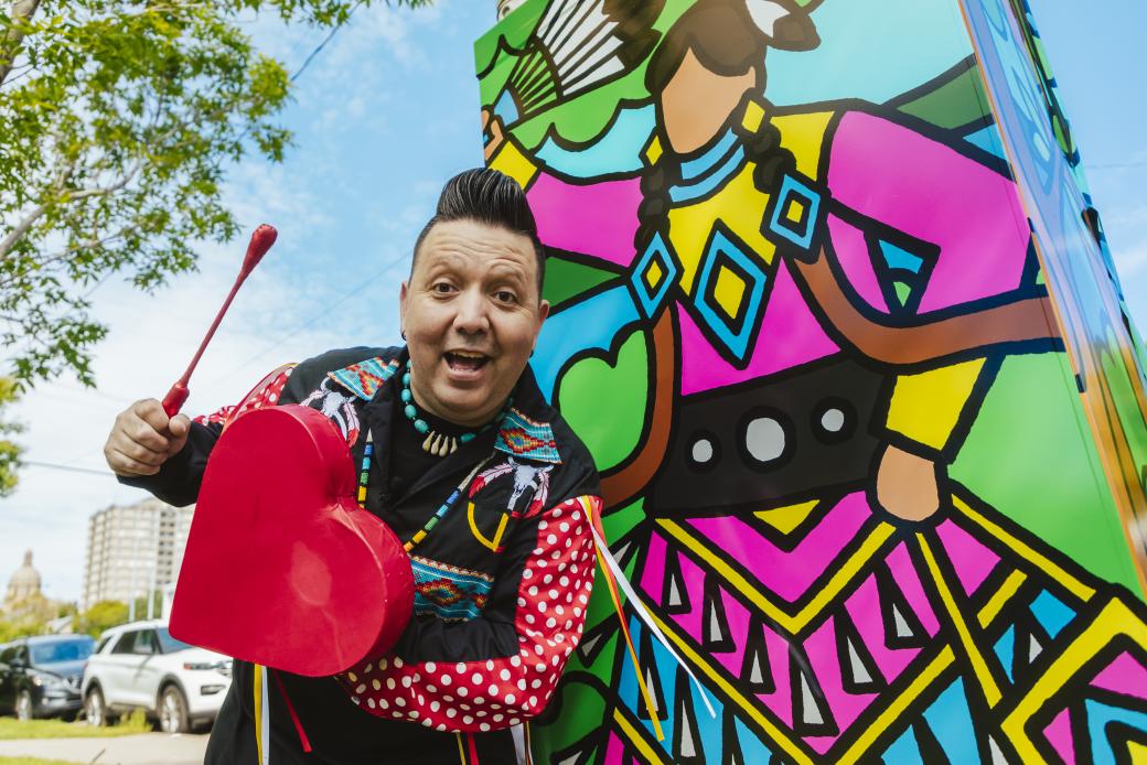 An Indigenous man holding a red stick and heart-shaped drum stands next to a traffic control box wrapped with his artwork of a jingle dress dancer holding a fan of feathers