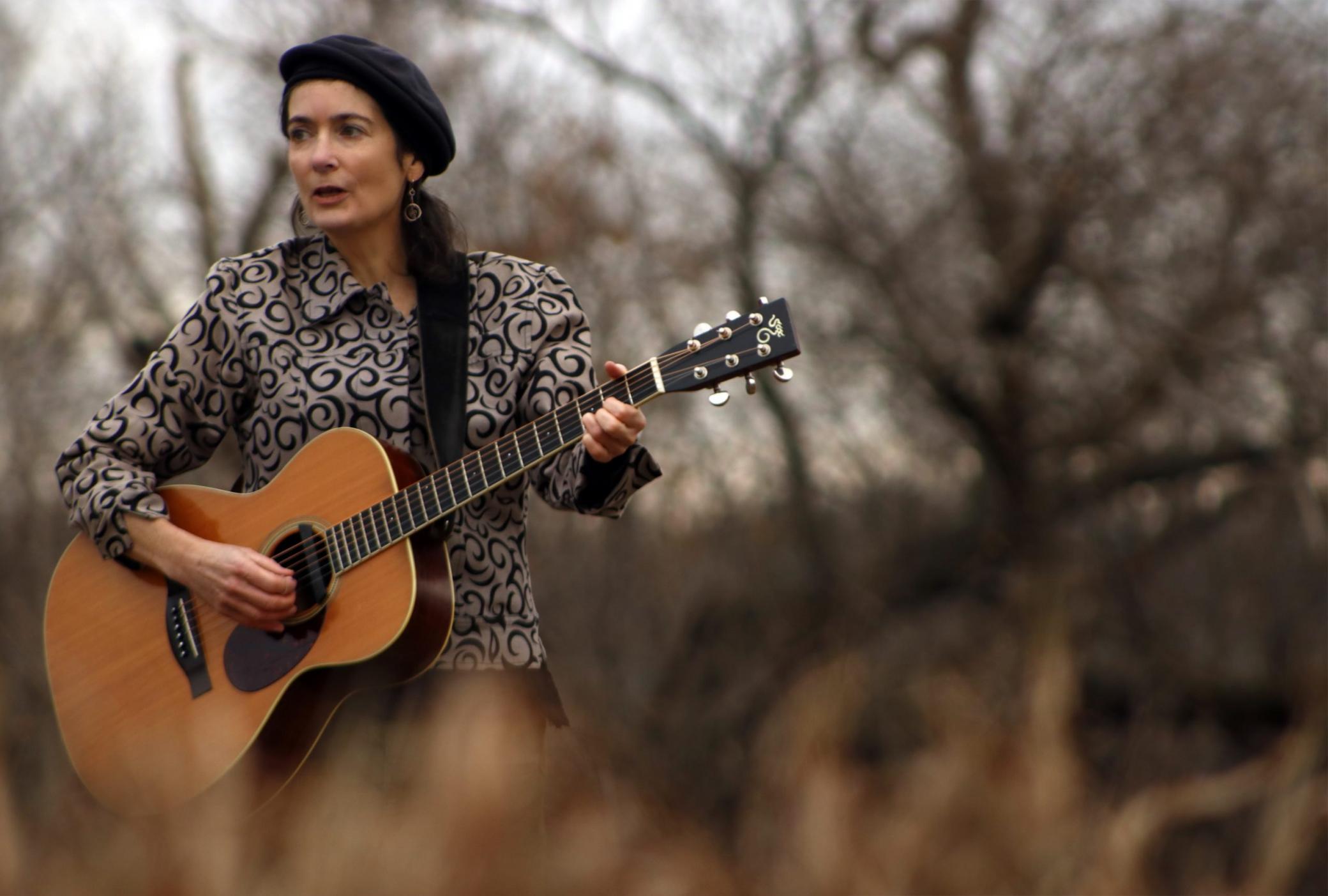 A woman, wearing a beret, plays an acoustic guitar outdoors, surrounded by trees. 