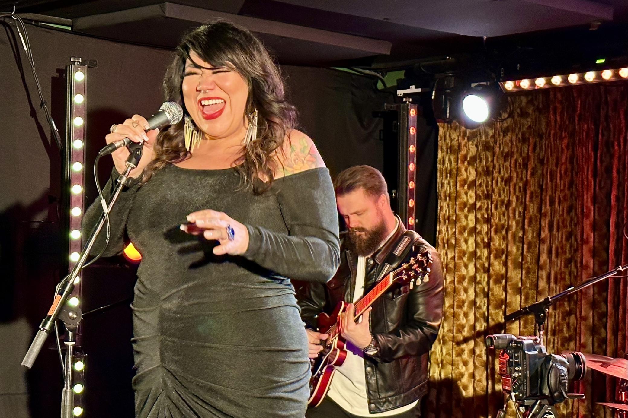  A woman in a black dress sings into a microphone as a bearded man plays guitar behind her on stage in a basement venue. 
