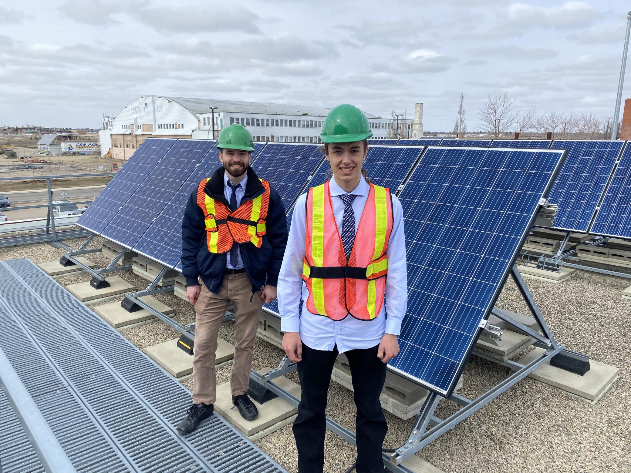 NAIT students Jack Harding and Ethan Buchanan standing in safety gear in front of the solar panels on a roof.