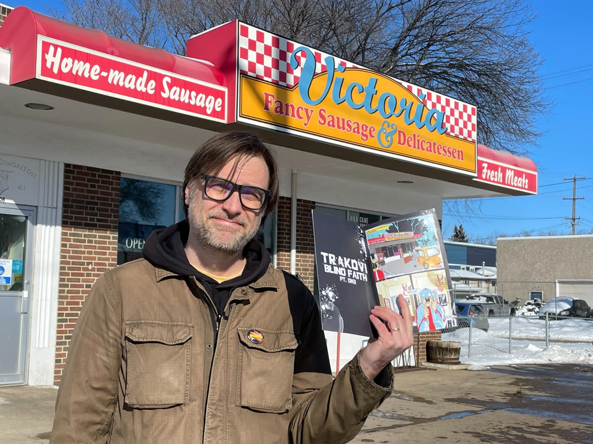 Adriean Koleric holds up a copy of his comic book, Trakovi: The Slav With No Remorse, in front of a deli shop in northeast Edmonton.
