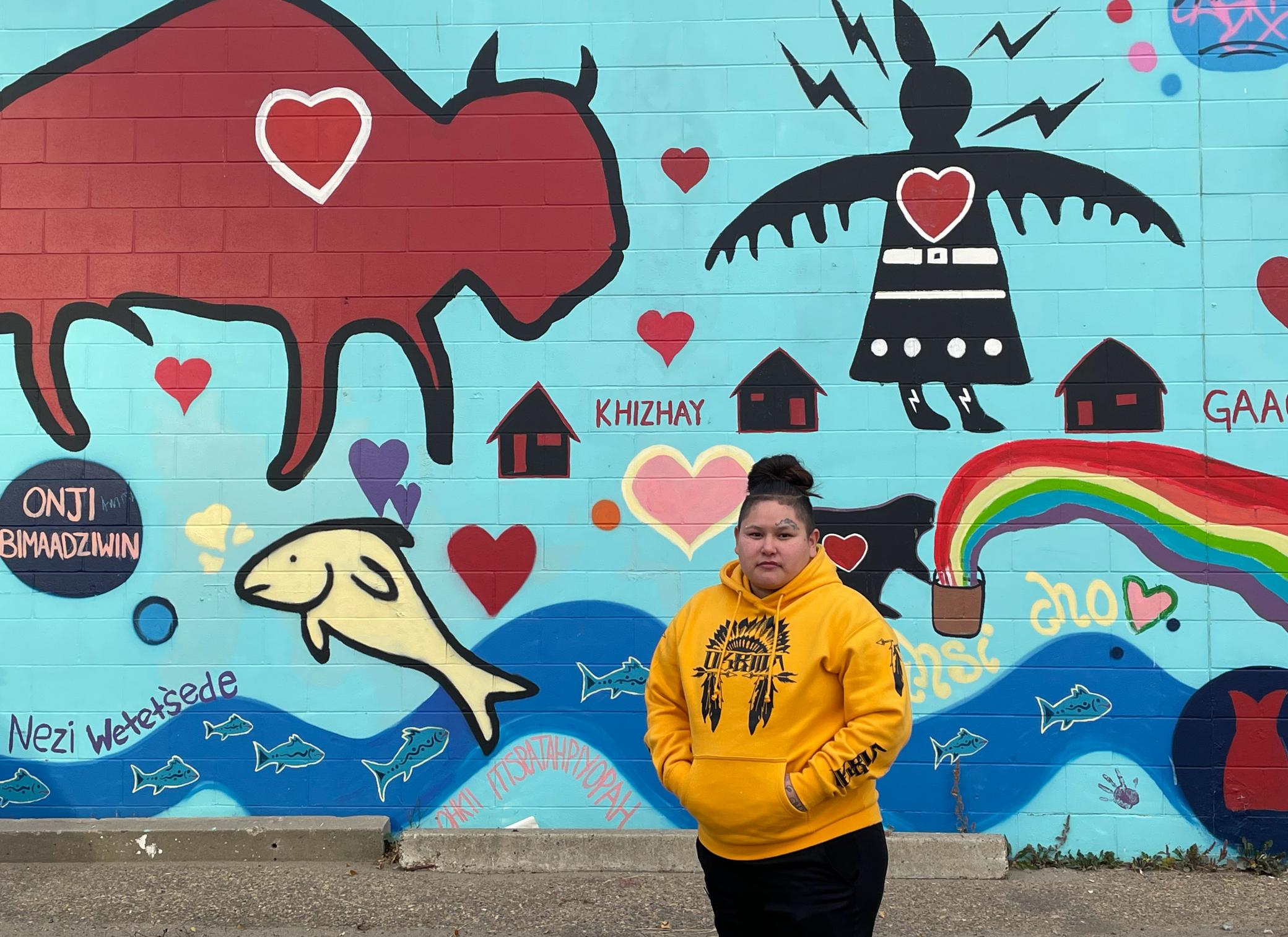 A woman in a yellow hoodie and black pants stands in front of a painted mural featuring hearts and Indigenous words and symbols, such as a buffalo. 