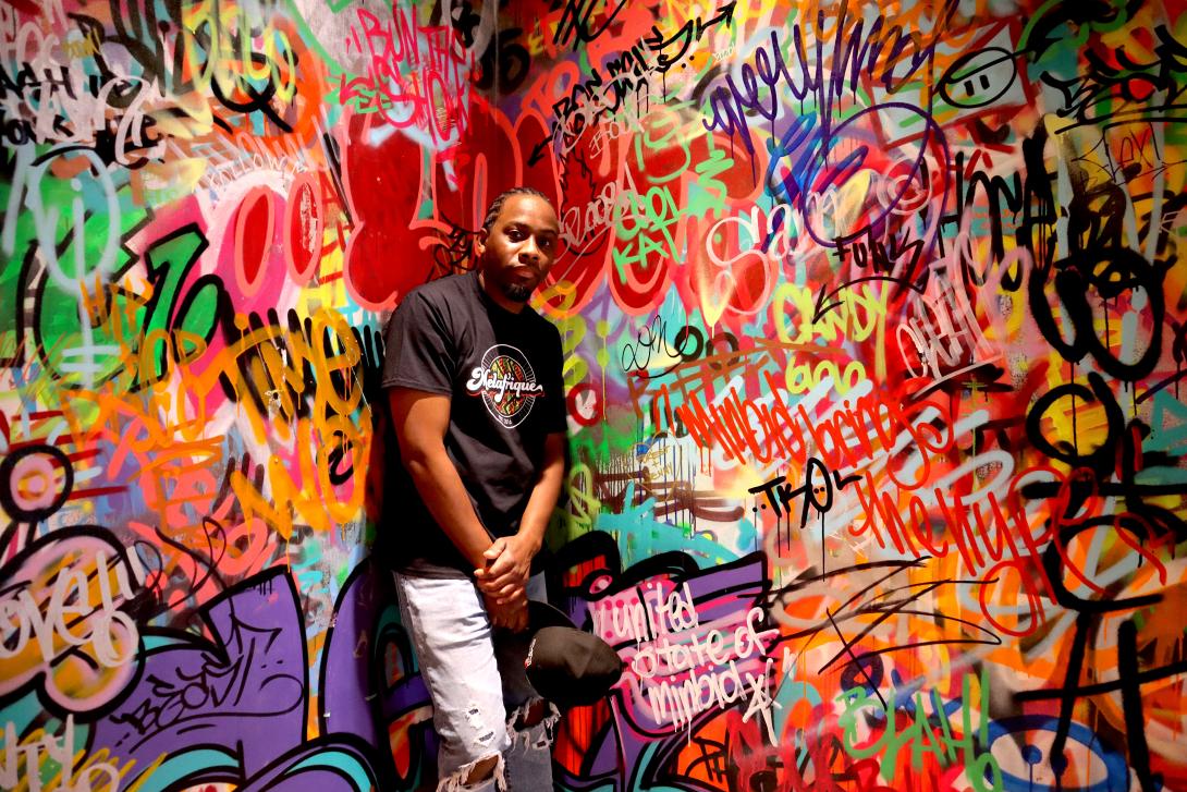 A black man stands in the corner with colourful graffiti covering the walls