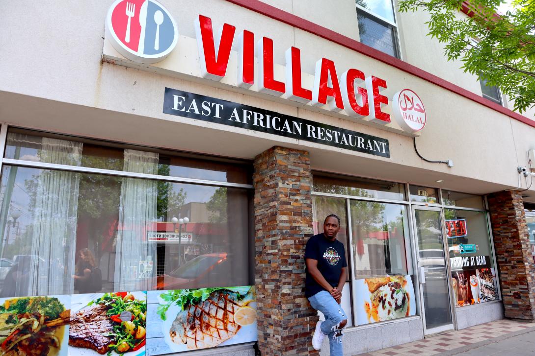 A black man leans against a pillar outside a restaurant. 
