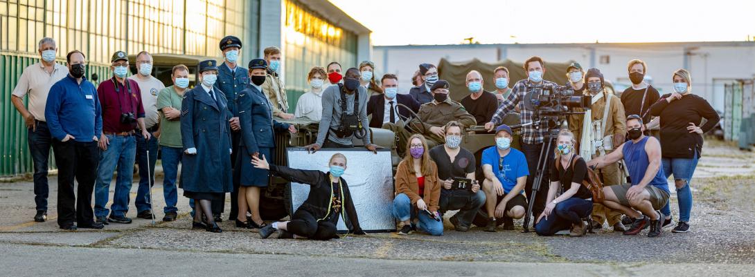A group of approx. 30 people, all in masks and some in costumes, stand outside the Blatchford hangar. 
