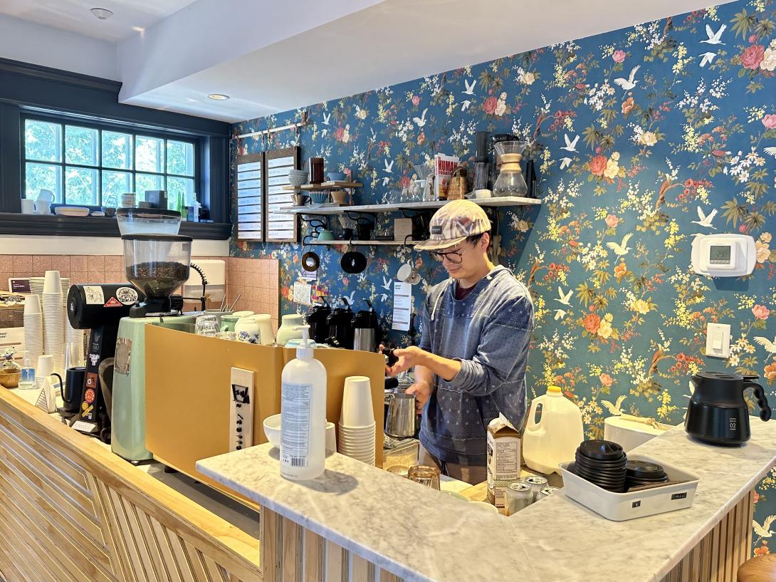 A man with a baseball cap makes a beverage behind the counter of a cafe. 