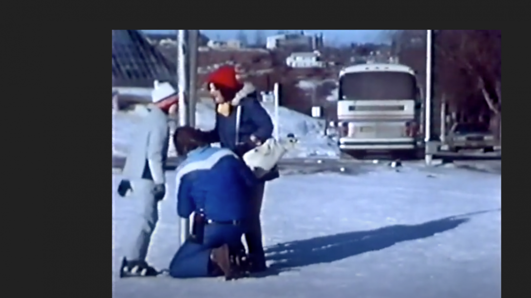 Screen shot of a child with his tongue stuck to a pole behind helped by 2 adults, all dressed in 80s snow gear. 