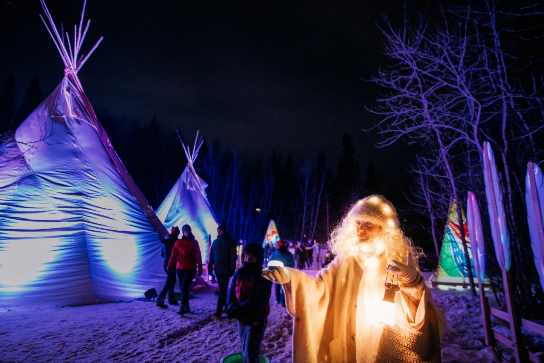 A festival performer dressed as a winter wizard and guests at the Indigenous camp at night. 