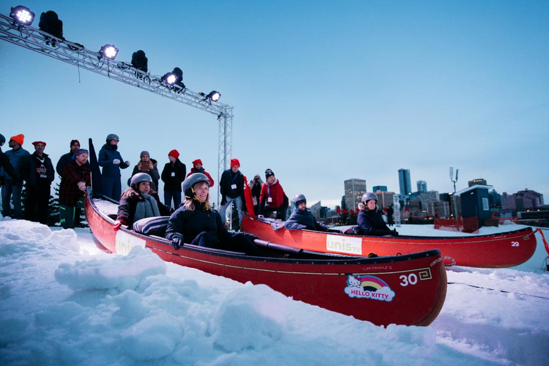 Two teams of racers sit in red canoes at the start of the ski hill course. 