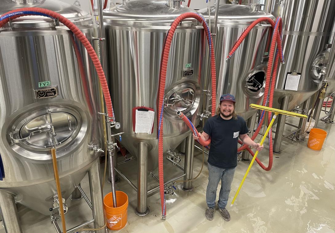A man holds a hose and a large squeegee as he stands in front of three silver vessels where beer is fermented. 