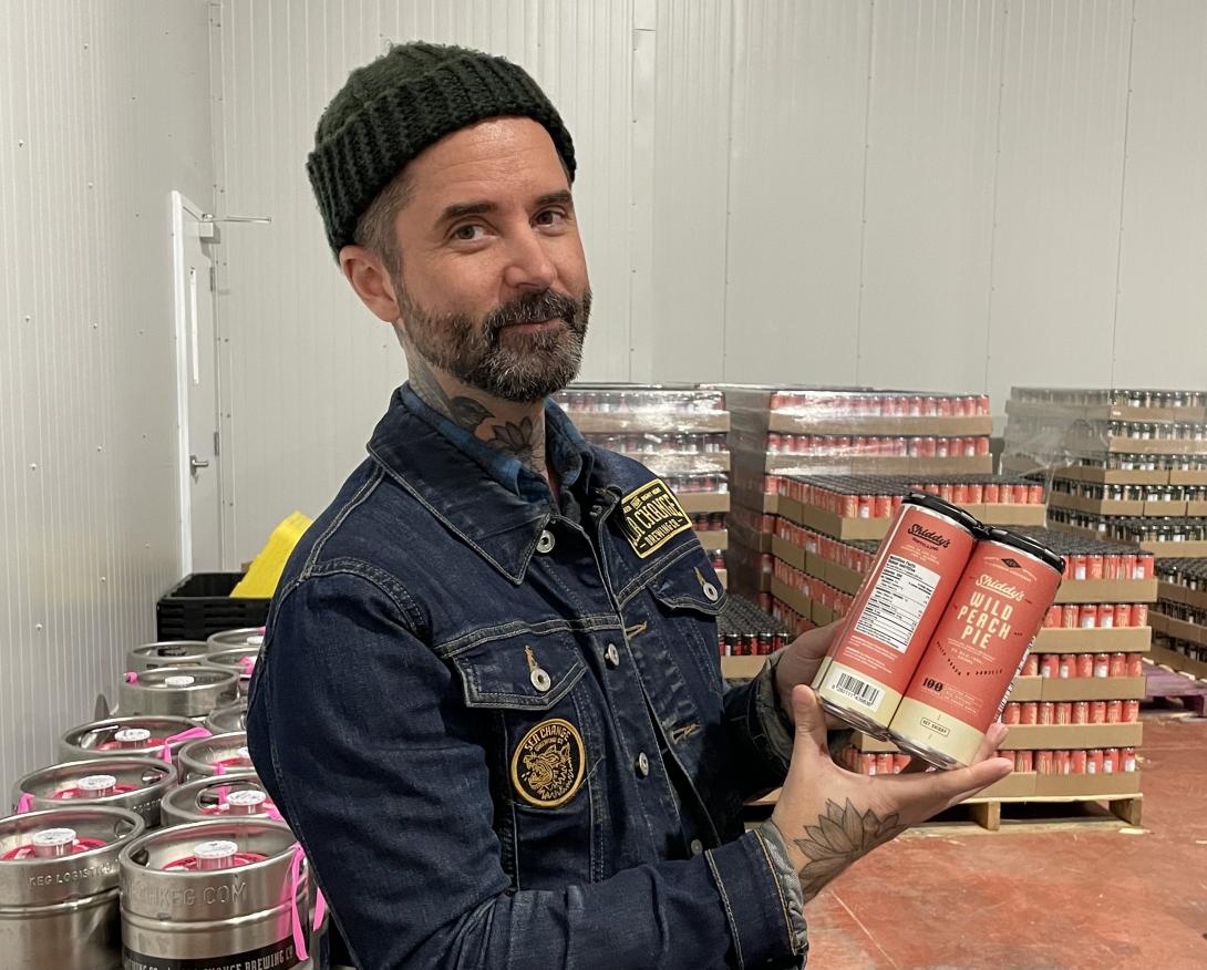 A man in a jean jacket and black beanie holds up a four-pack of cans in a storage room of kegs and flats of beer.