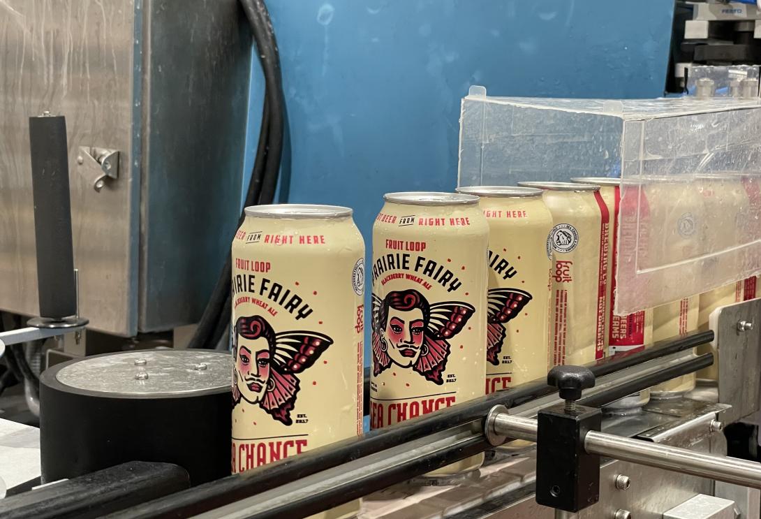 A line of cans of Sea Change’s Prairie Fairy beer stand in a line on a conveyor belt, waiting to be moved and packaged. 