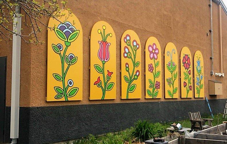 Six paintings of flowers are attached to a wall next to a community garden. 