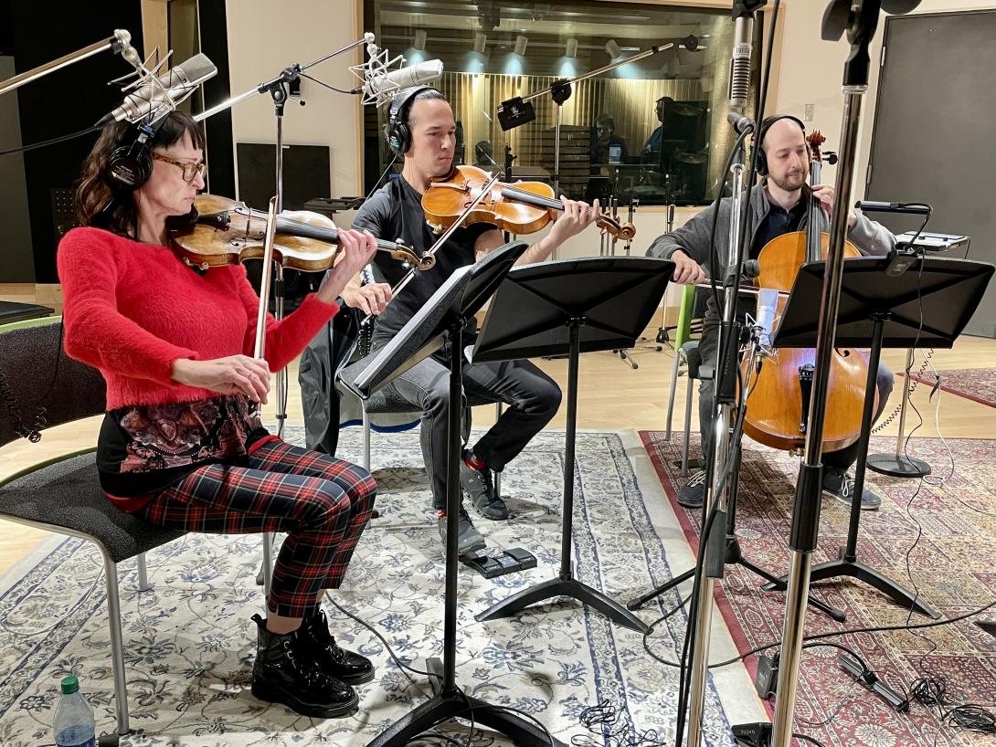 A violin player, a violist and a cellist read off music stands and play their instruments as they sit in a recording studio with rugs on the floor.