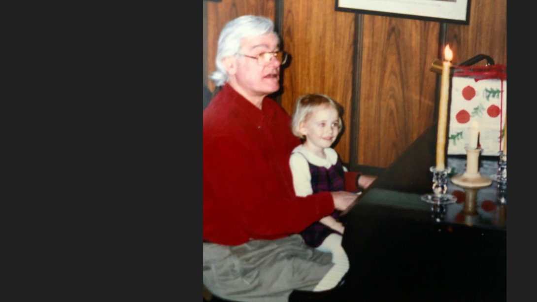A man with white hair and glasses plays a piano with his young granddaughter on his lap. 