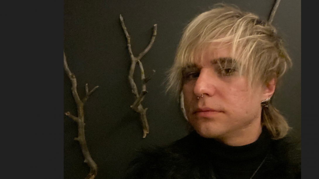 A close-up of a person with dyed blonde hair standing in front of some twigs attached to a wall. 
