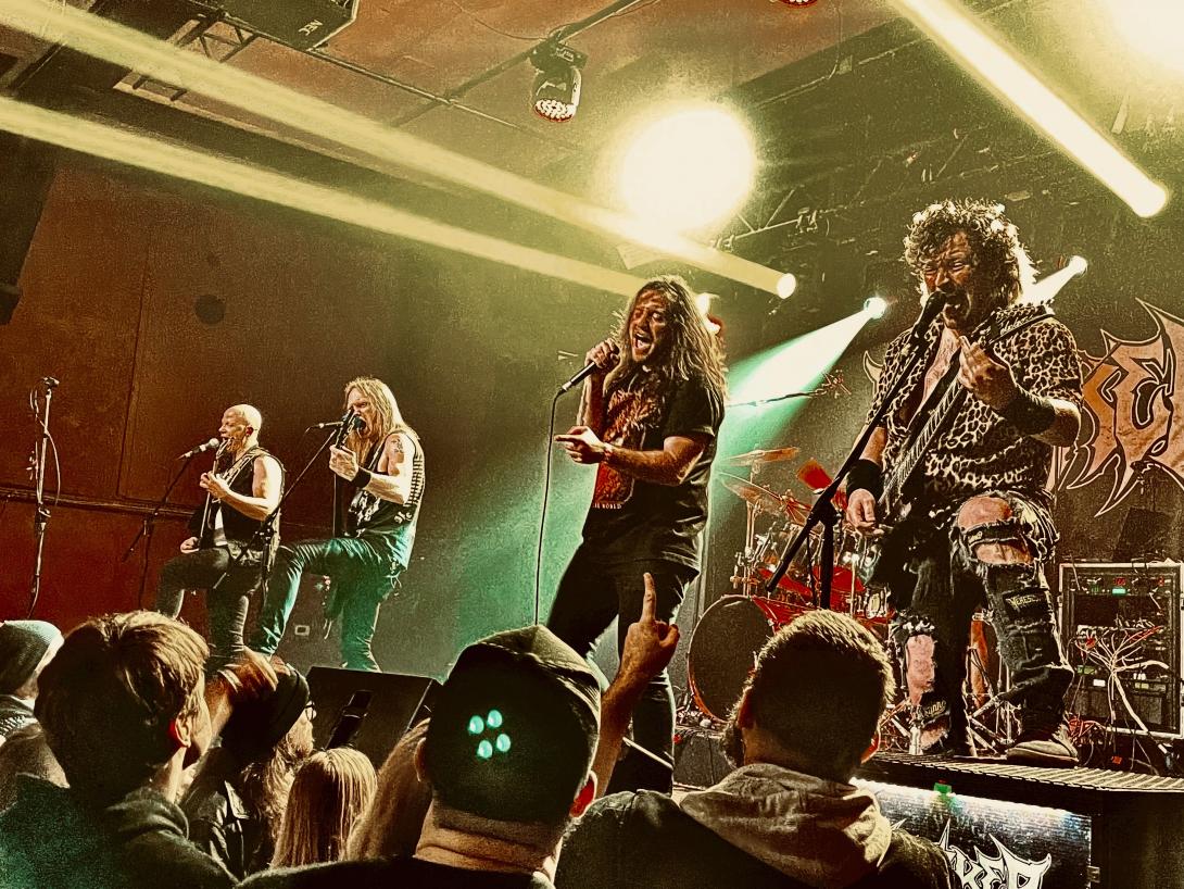  A group of five metal musicians perform on stage in front of some fans as yellow strobe lights stream over their heads. 