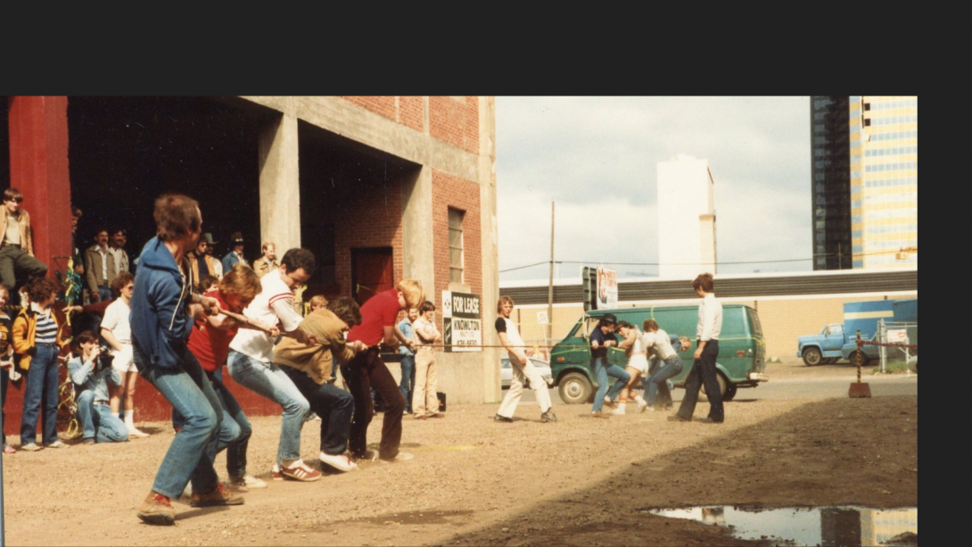 Two groups of men take part in a tug of war in a gravel alley next to a brick building. 