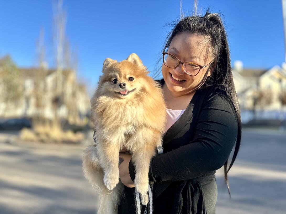 A smiling woman with glasses holds a smiling pomeranian with his tongue sticking out.