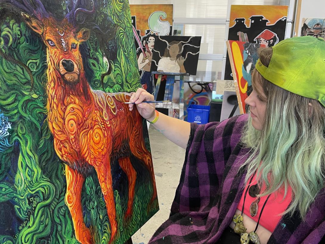 A person sits next to a canvas and puts the finishing touches on a fantastical orange deer.