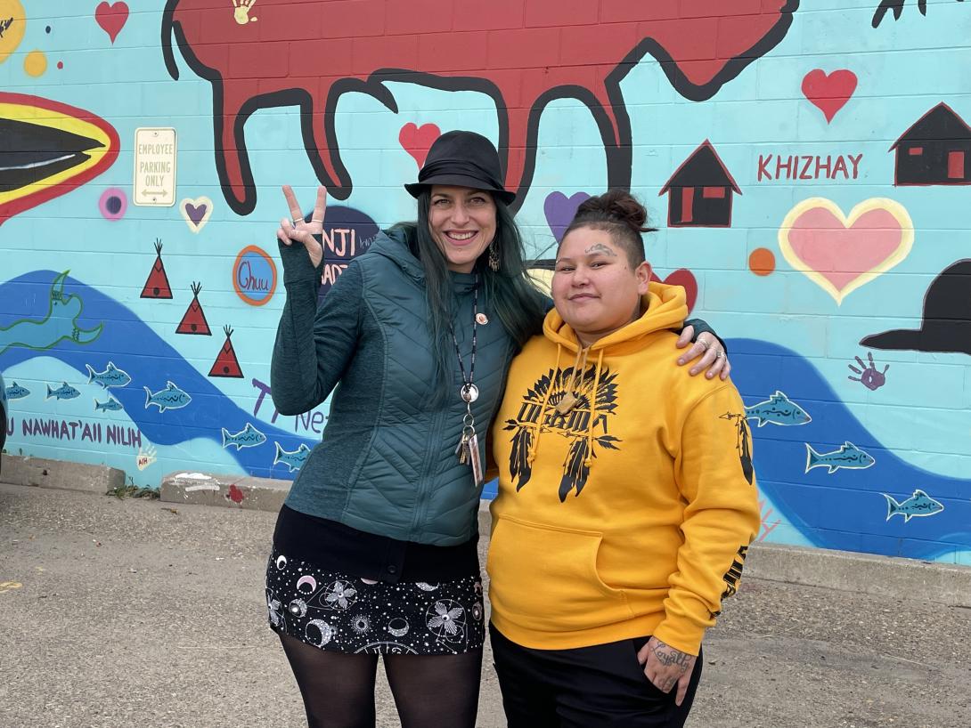 Two women stand in front of a colourful wall with Indigenous symbols and words.