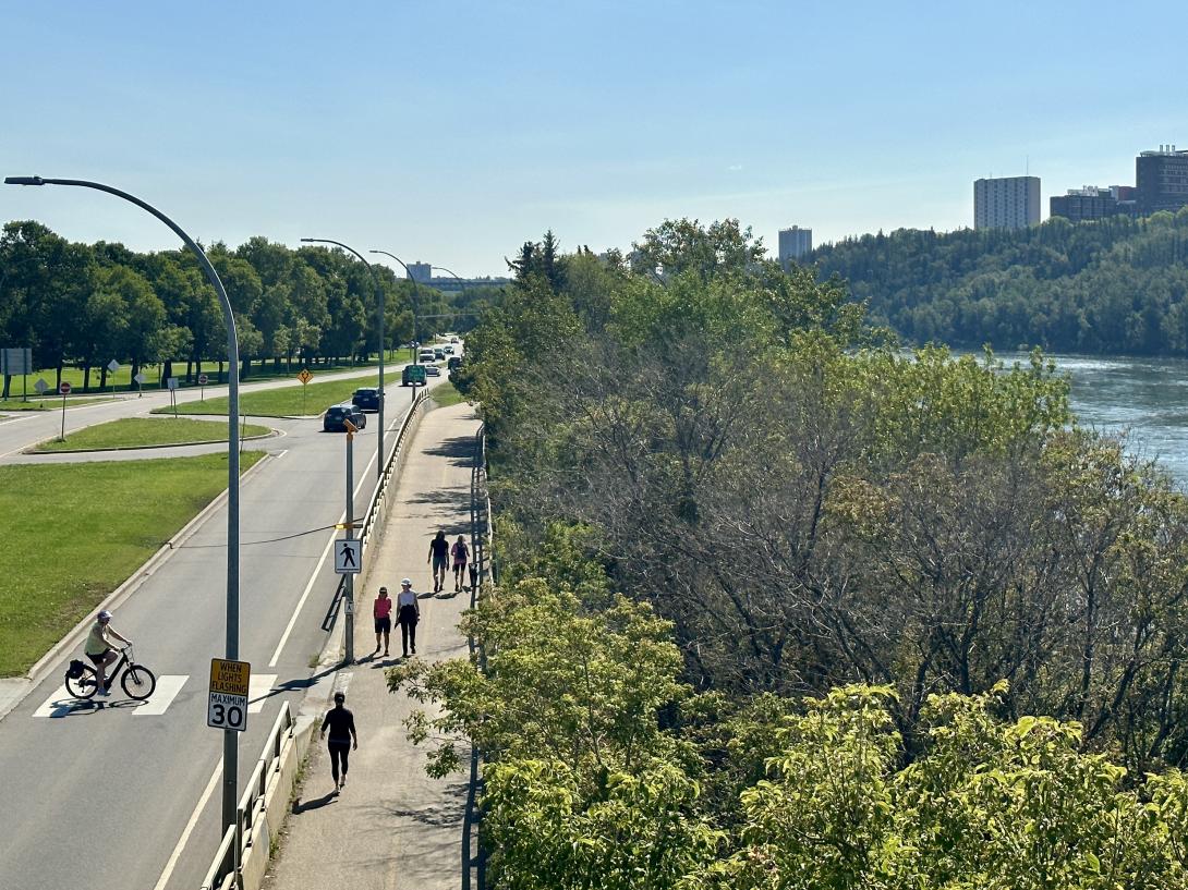 Groups of people walk along a paved path next to a road on their left and some trees and the North Saskatchewan River on their right.