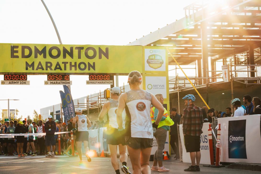Three people with their backs to the camera run toward an overhead sign that says Edmonton Marathon. 