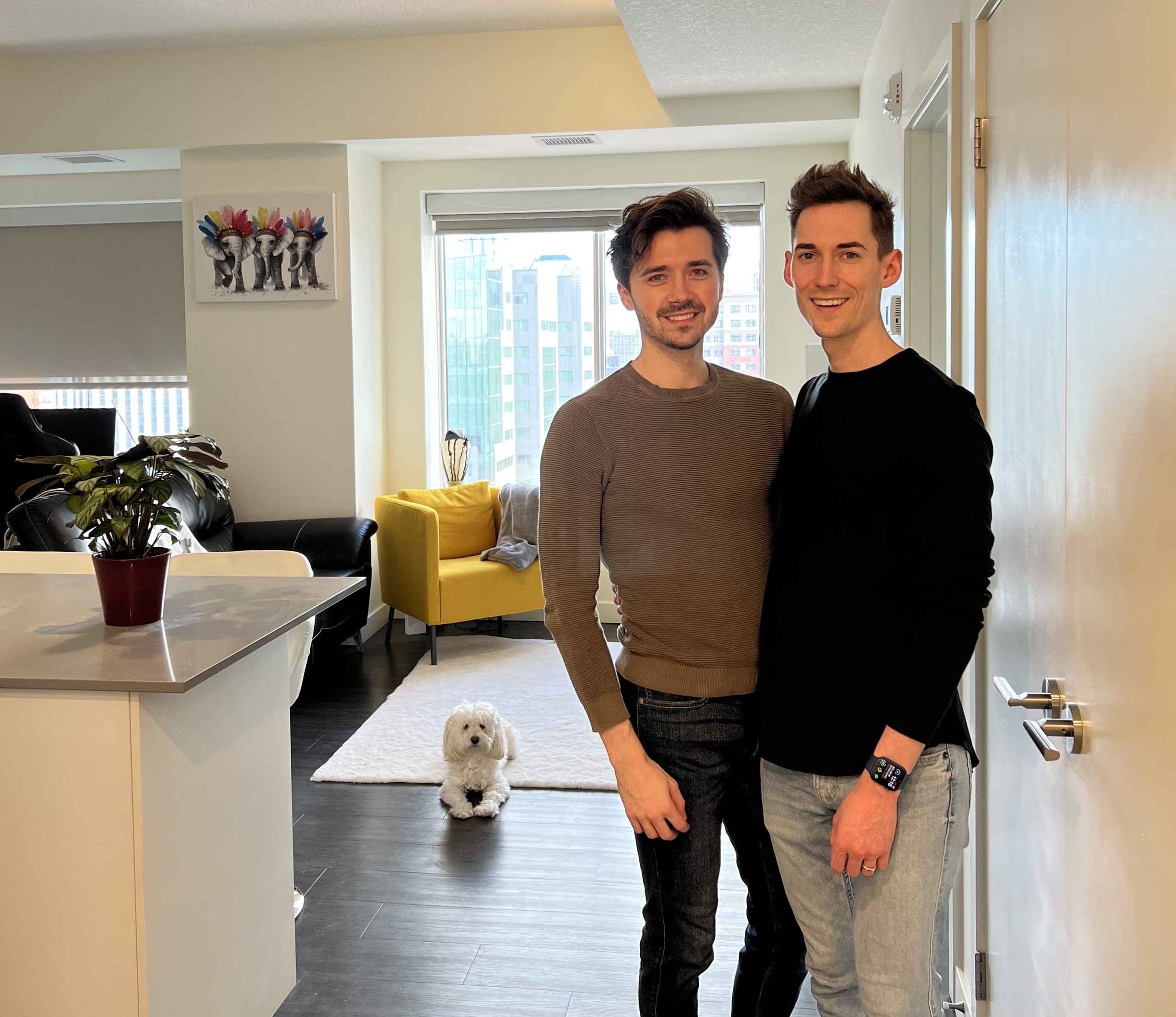 Two men standing in the doorway to their condo with a small white dog sitting on the floor next to them.