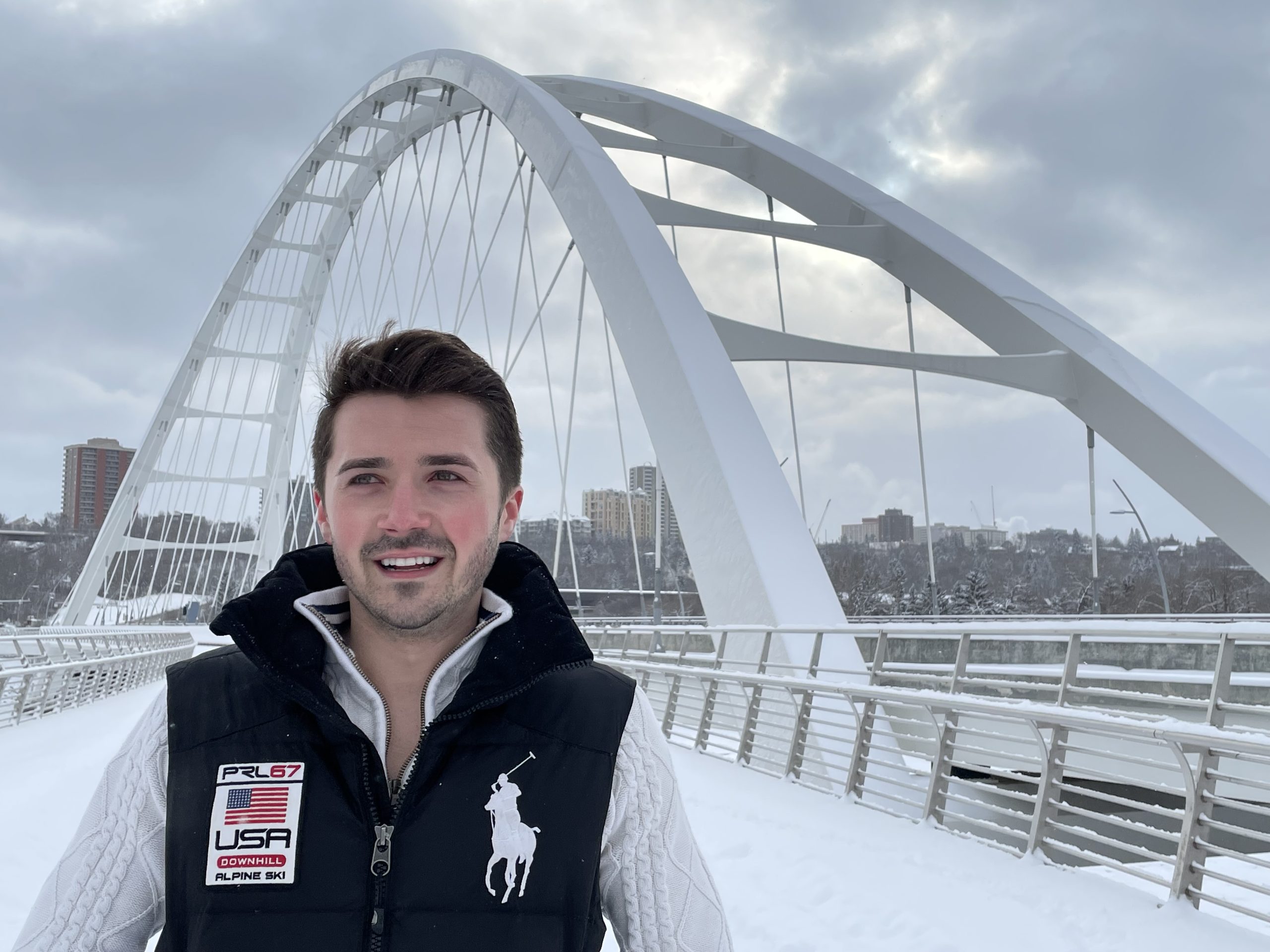 A young man in a vest standing outside in front of an arched bridge in winter.