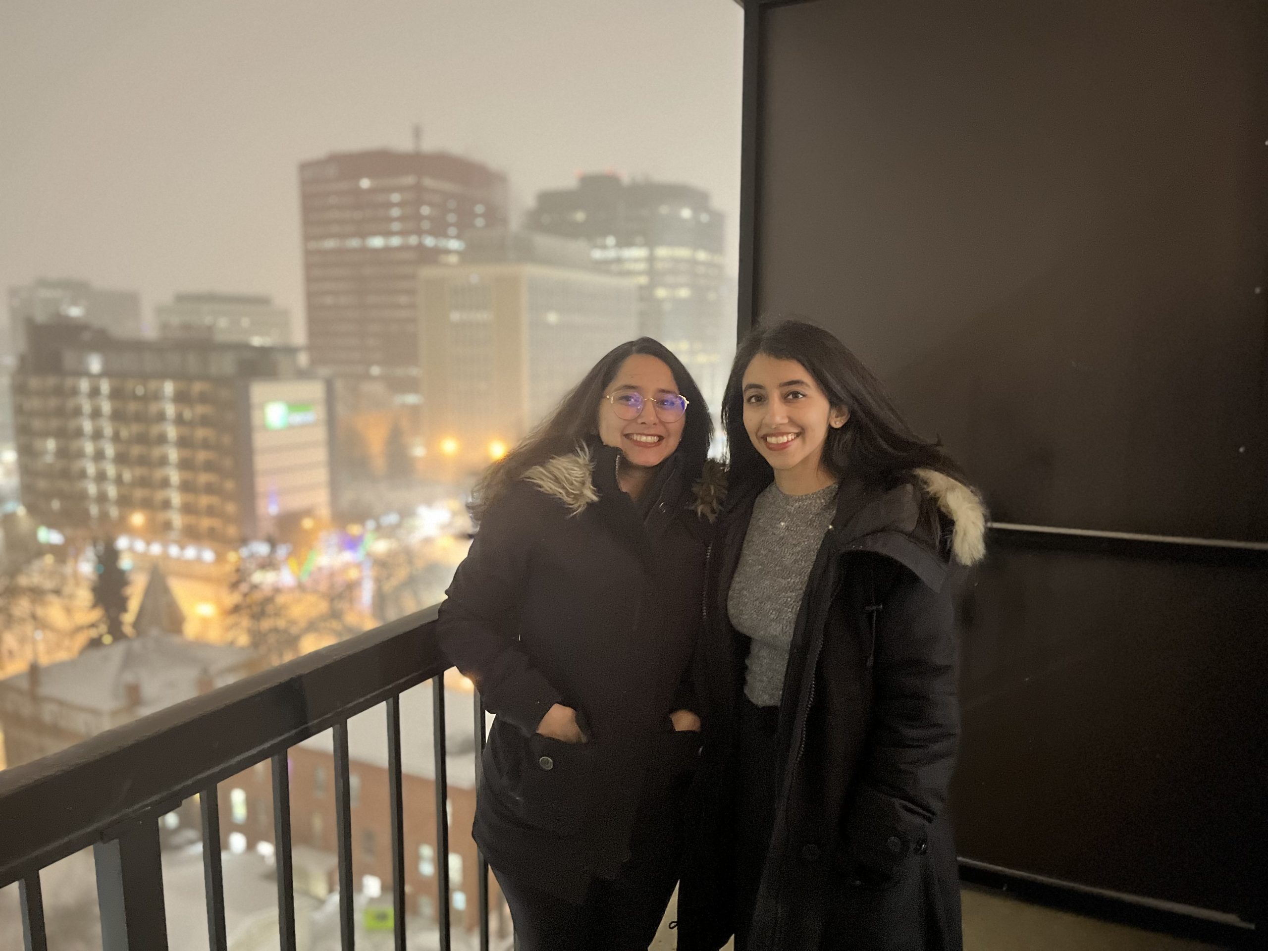 Two young women stand outside on a balcony on a winter night.