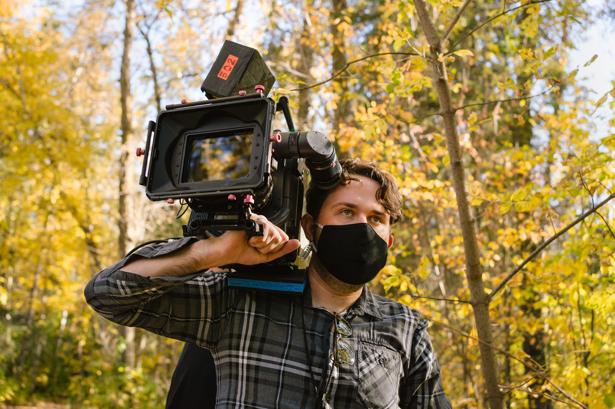 A man carrying a professional film camera & wearing a mask.