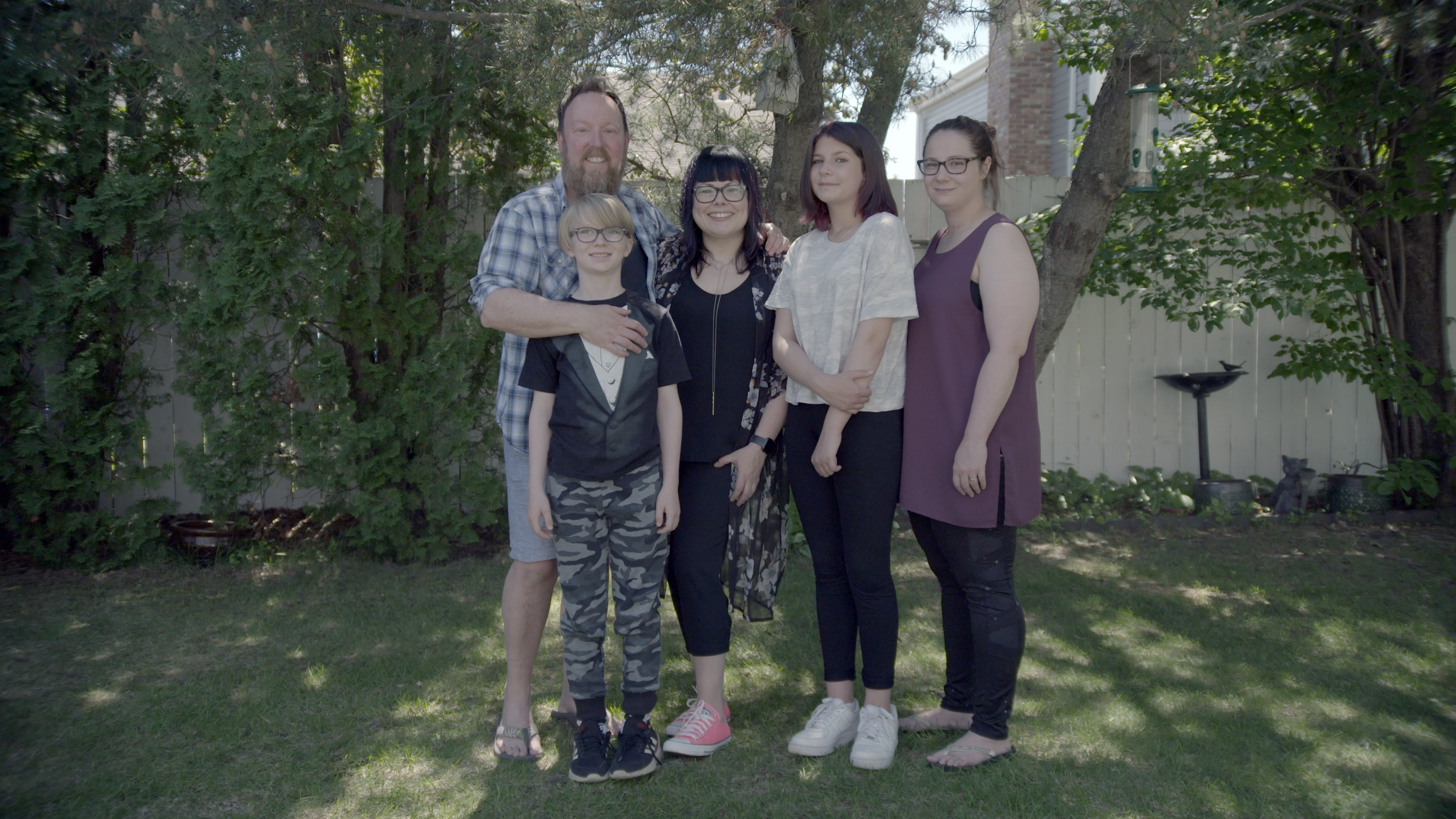 A family of 5, parents with 3 children, stand outside in front of a tree.