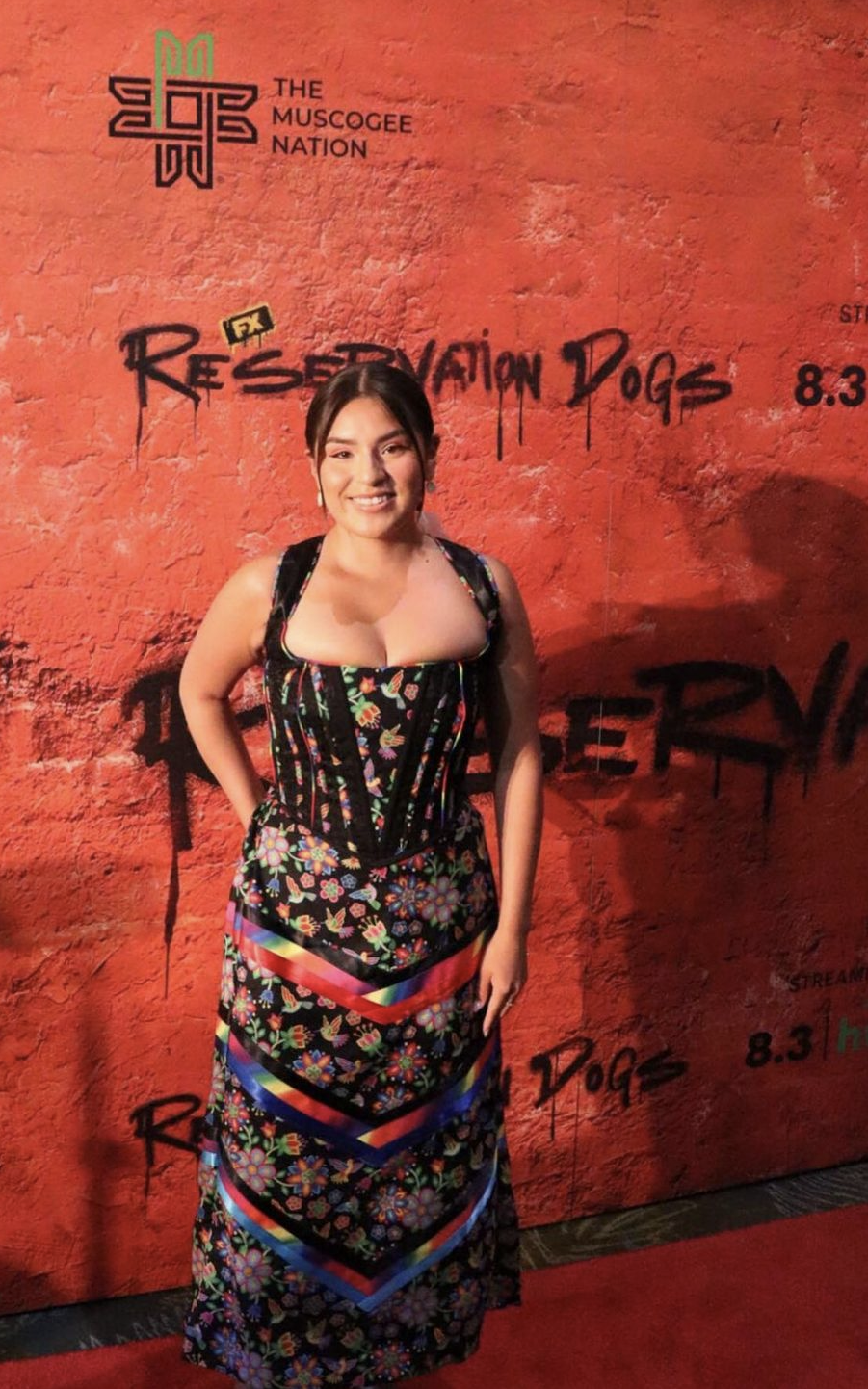 Paulina Alexis, wearing a colourful dress with floral patterns and geometric designs, stands in front of a red wall with "Reservation Dogs" written on it. She is smiling and posing for the camera.