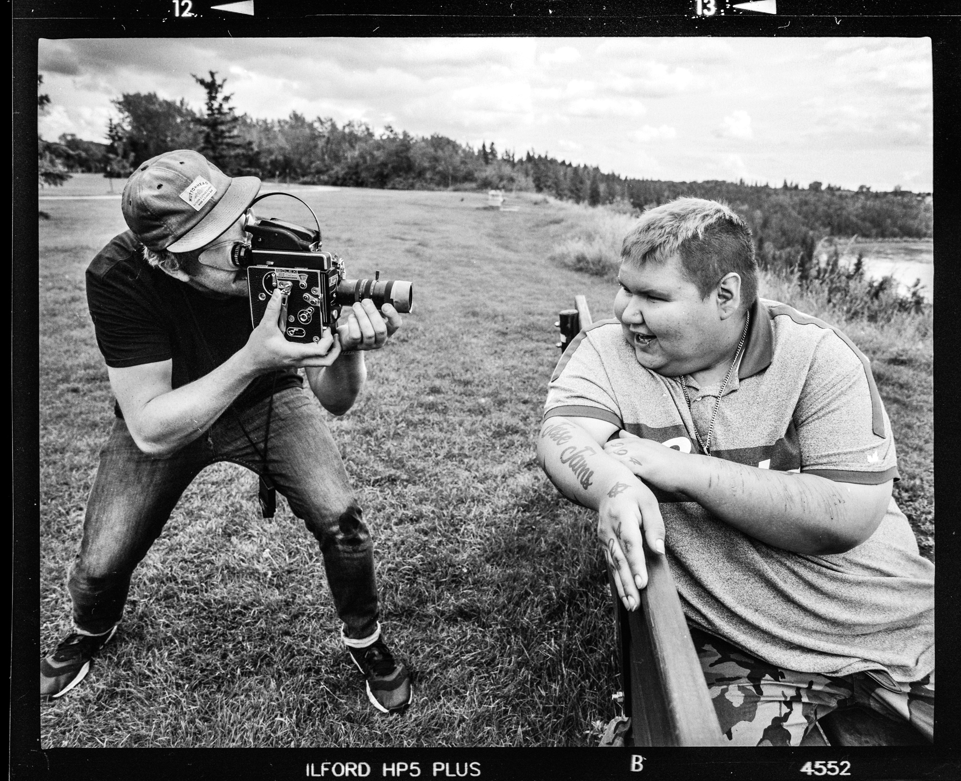 A man behind a video camera taking close of shots of another man sitting on a bench in black and white.