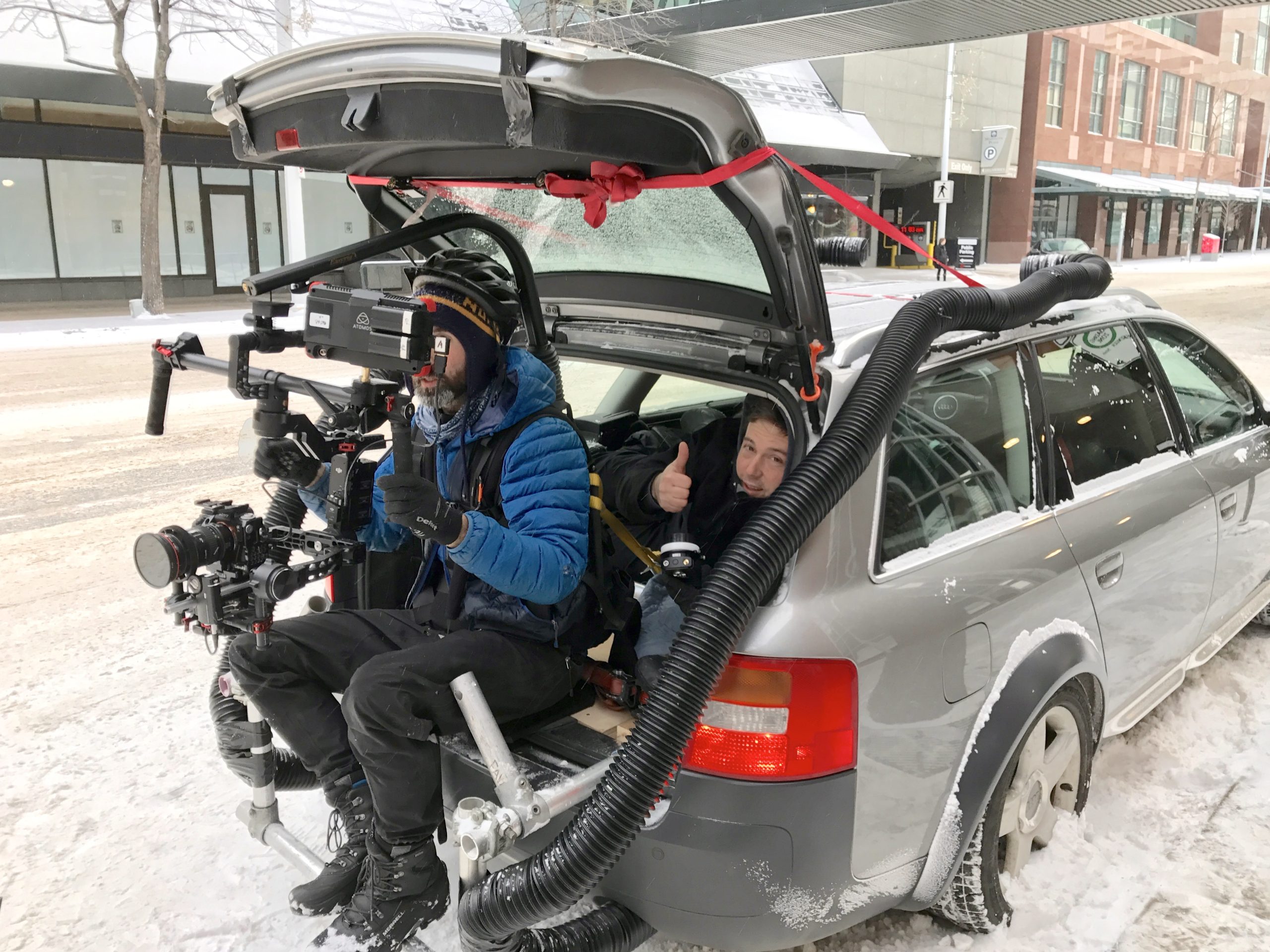 A film crew riding in the back of an open hatchback on a snowy street.