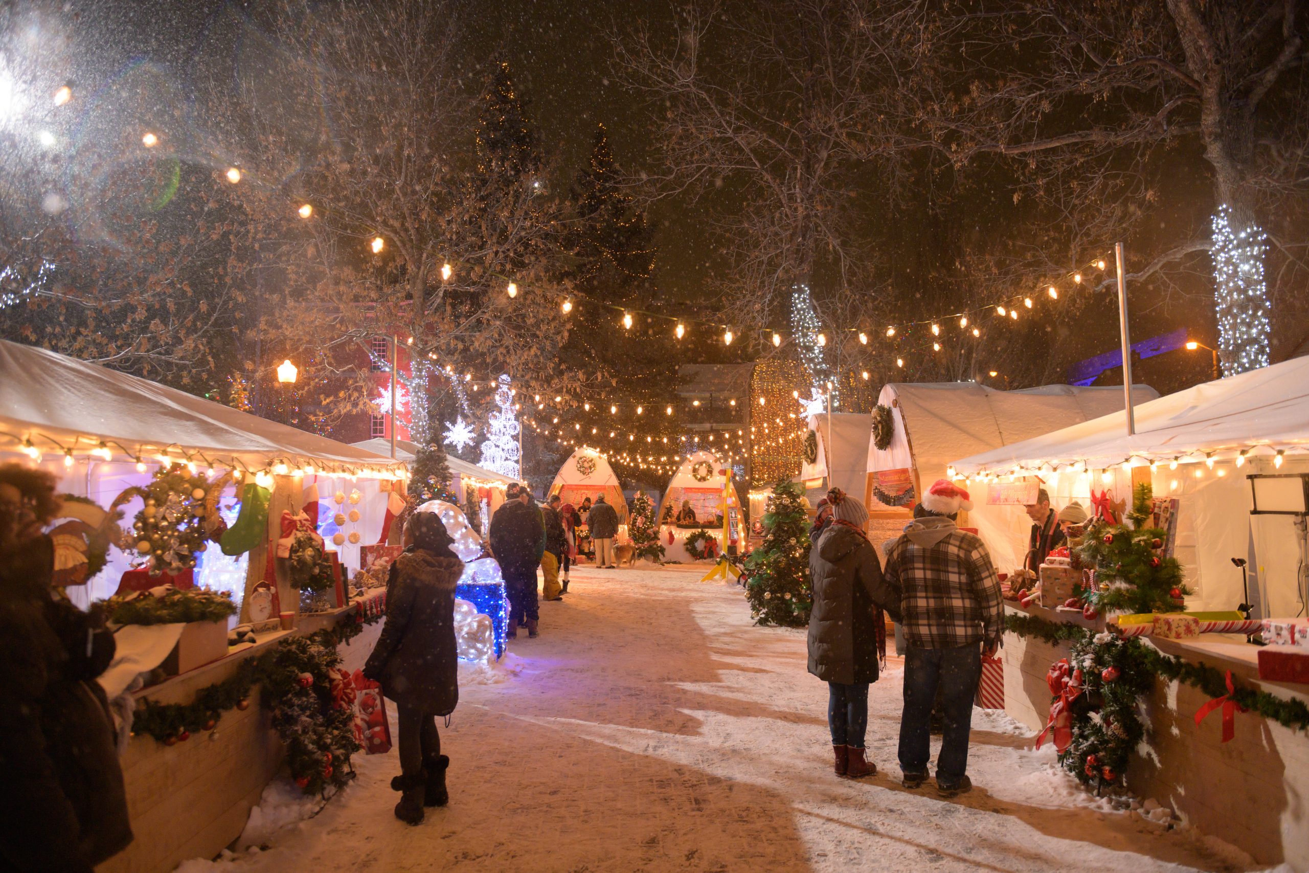 A movie set with guests enjoying a Christmas market with string lights in the snow.