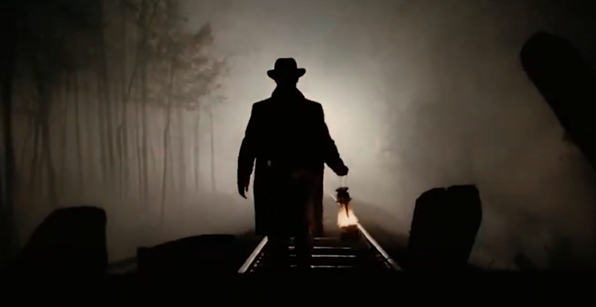 The shadow on a man in a trench coat and hat carrying a lantern walking down railway tracks at night.