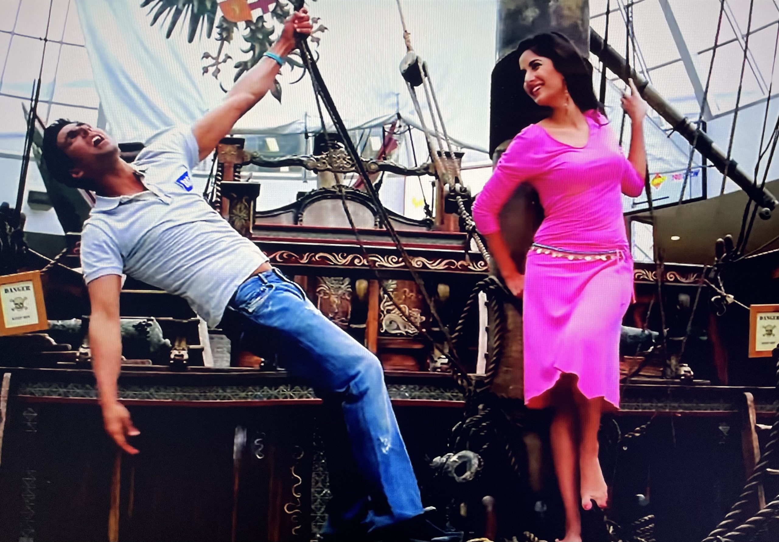 An actress in a pink dress and an actor in a white shirt and jeans perform in a scene on a wooden boat.