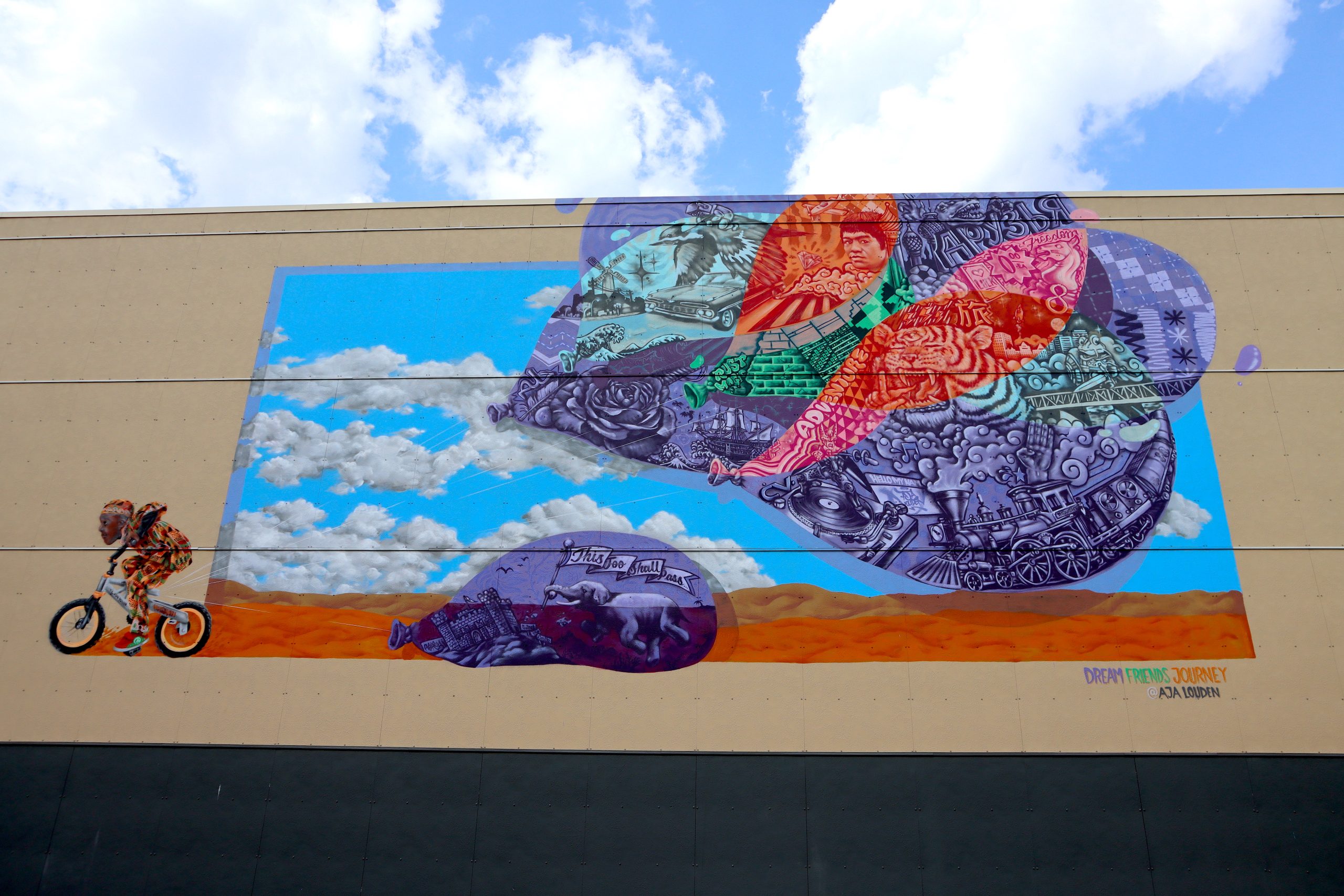 A mural with a person riding a bike to the left and giant colorful balloons filled with diverse images including a tiger, train and flowers., Muralist AJA Louden painted Dream Friends Journey, which includes images of Bruce Lee and a turntable, in 2018.