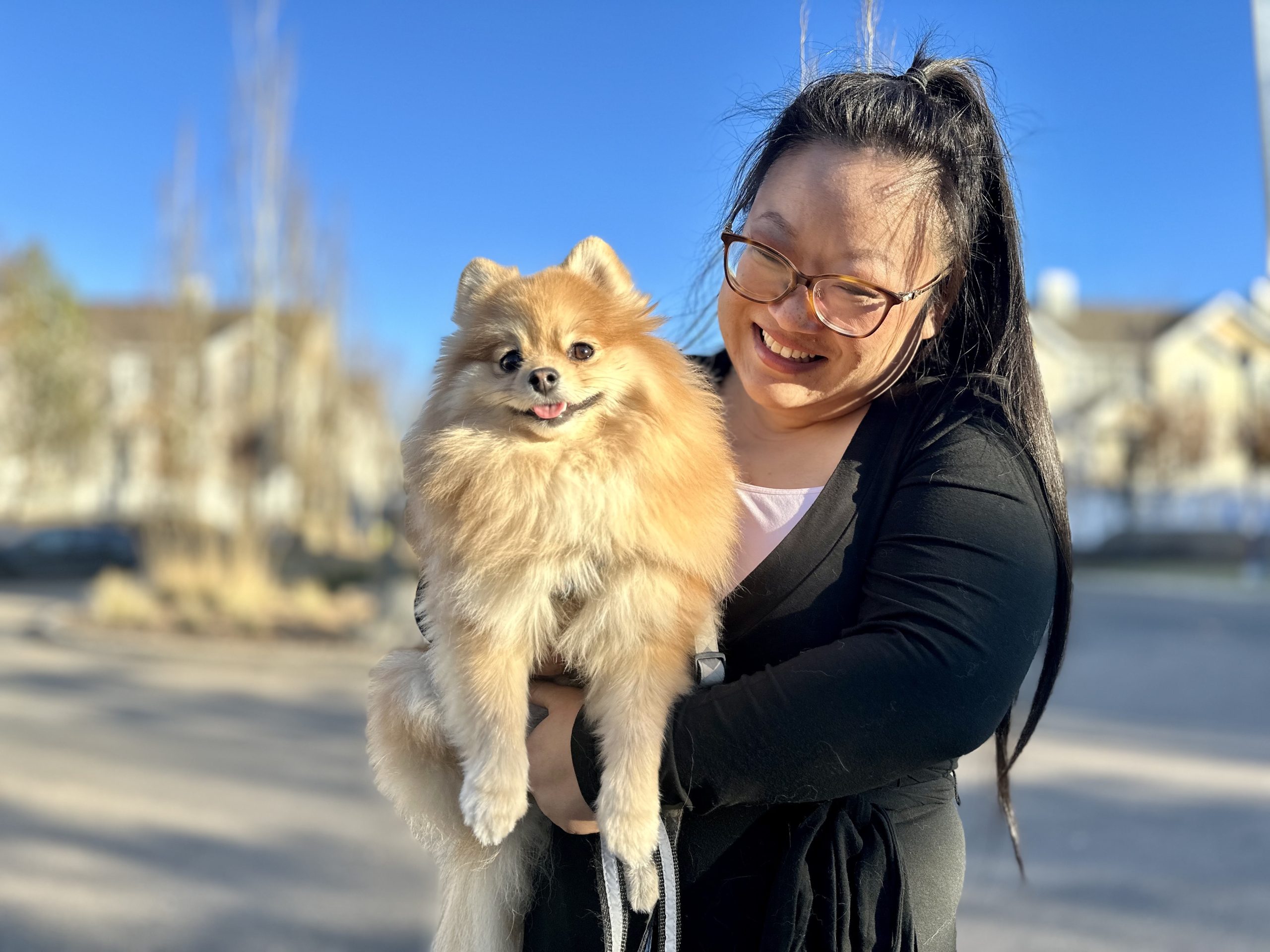 A smiling woman with glasses holds a grinning pomeranian with his tongue sticking out.