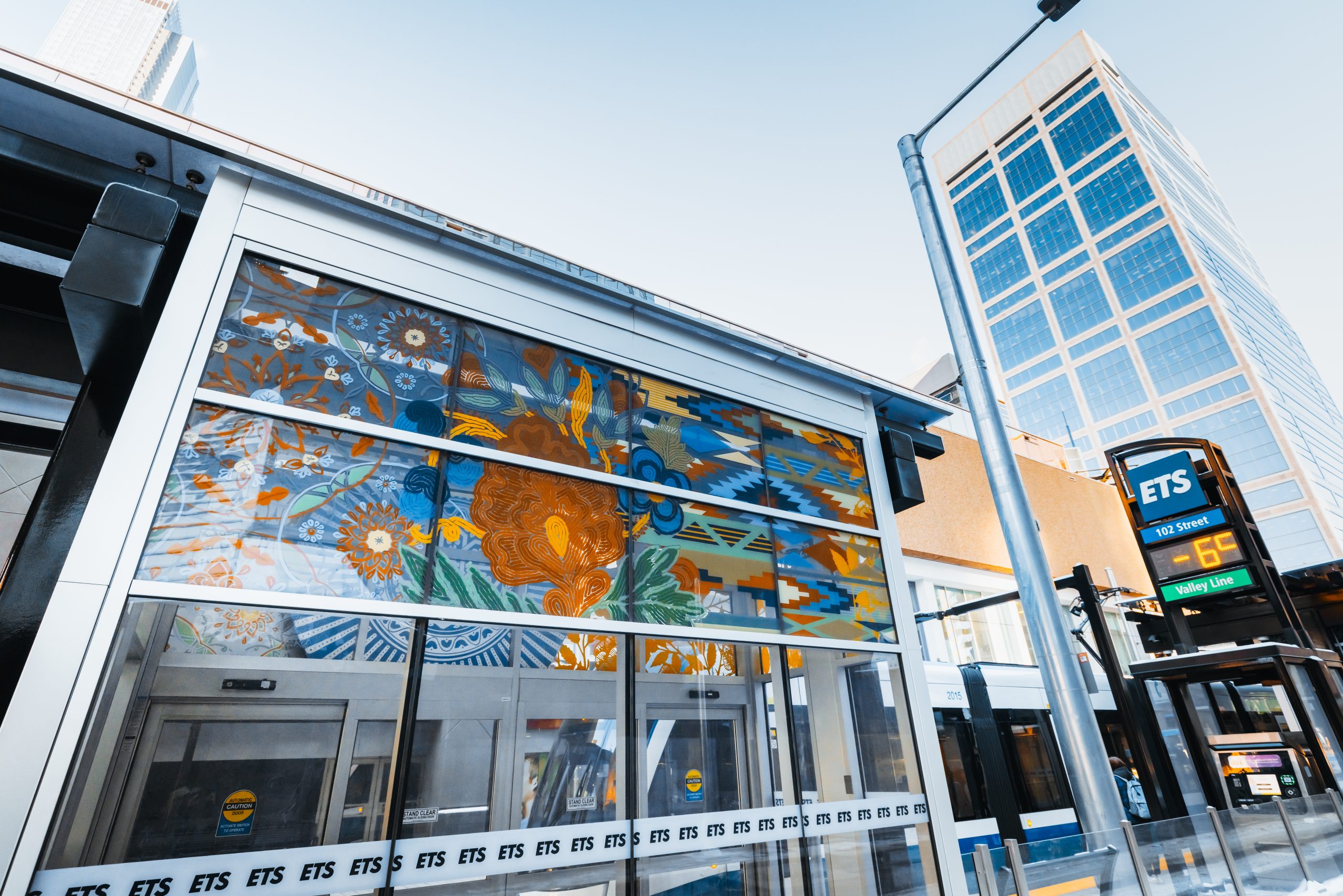 A glass LRT shelter is decorated with art of flowers and various striped patterns.